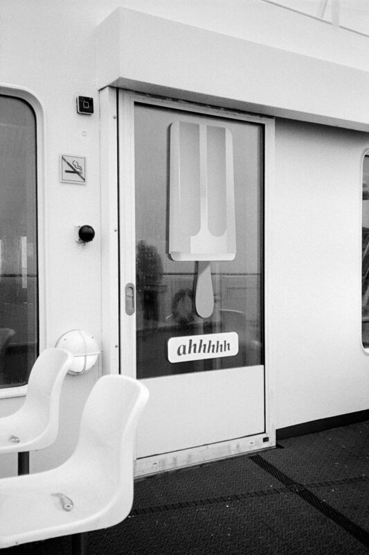 B&W film photo of a ferry door featuring a popsicle graphic and "ahhhhh" text. Grainy texture, white chairs in foreground.