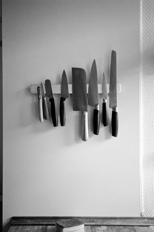 Grainy B&W shot of various kitchen knives aligned on a magnetic wall strip. High contrast with minimalist, domestic textures.