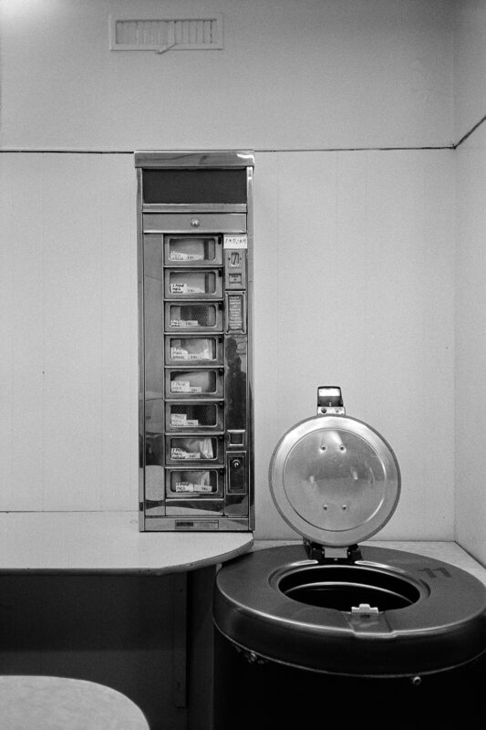 Black-and-white footage of a vintage vending machine next to a large spin dryer in a laundromat. A minimalist composition in shades of gray.