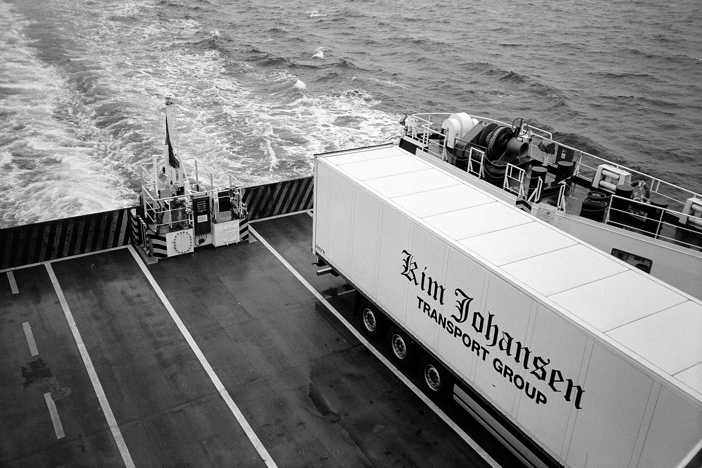 B&W high-angle shot of a Kim Johansen truck on a ferry deck. Grainy textures of the churning sea wake and industrial deck.