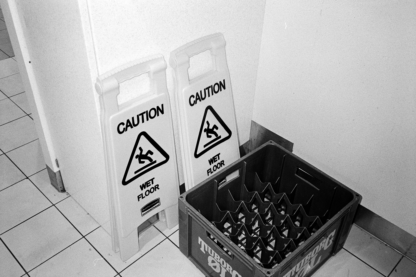 Grainy B&W photo of "Caution Wet Floor" signs next to a Tuborg beer crate. Ferry restaurant scene with high contrast film grain.