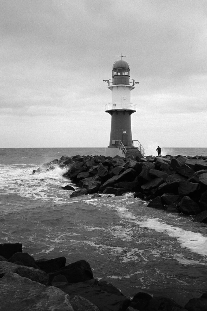 B&W film photo of a lighthouse on a rocky pier with crashing waves under a moody, overcast sky in Warnemünde.