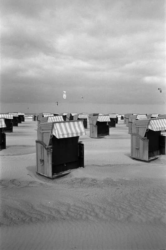Grainy B&W shot of striped beach chairs on a sandy shore under a cloudy sky, with kites visible in the distance.