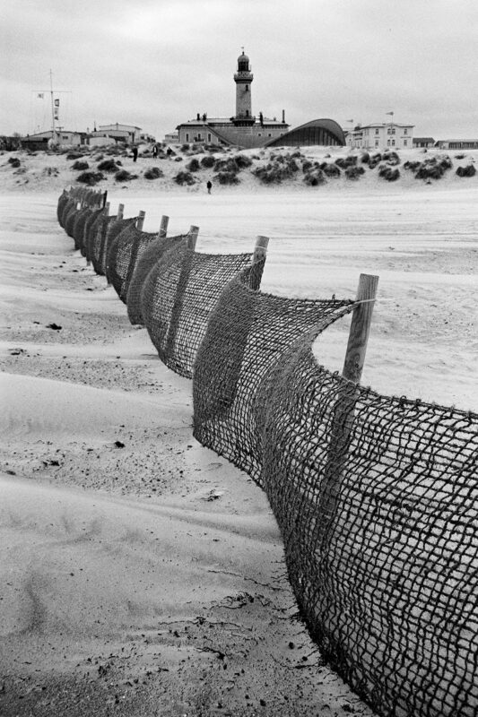 Grainy B&W shot of a mesh sand fence leading the eye toward a distant lighthouse and coastal buildings in Warnemünde.