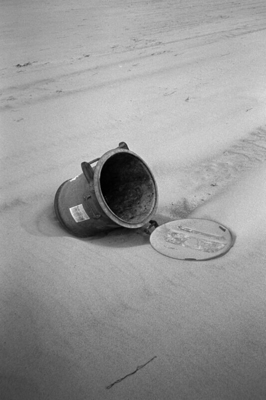 B&W close-up of a weathered metal trash can lying on its side in the sand, showing grainy textures and subtle details.