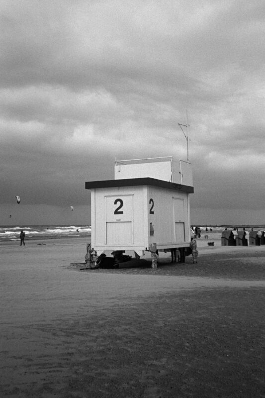 B&W film photo of a lifeguard station numbered '2' on a windy beach with kites flying over the rough Baltic Sea.