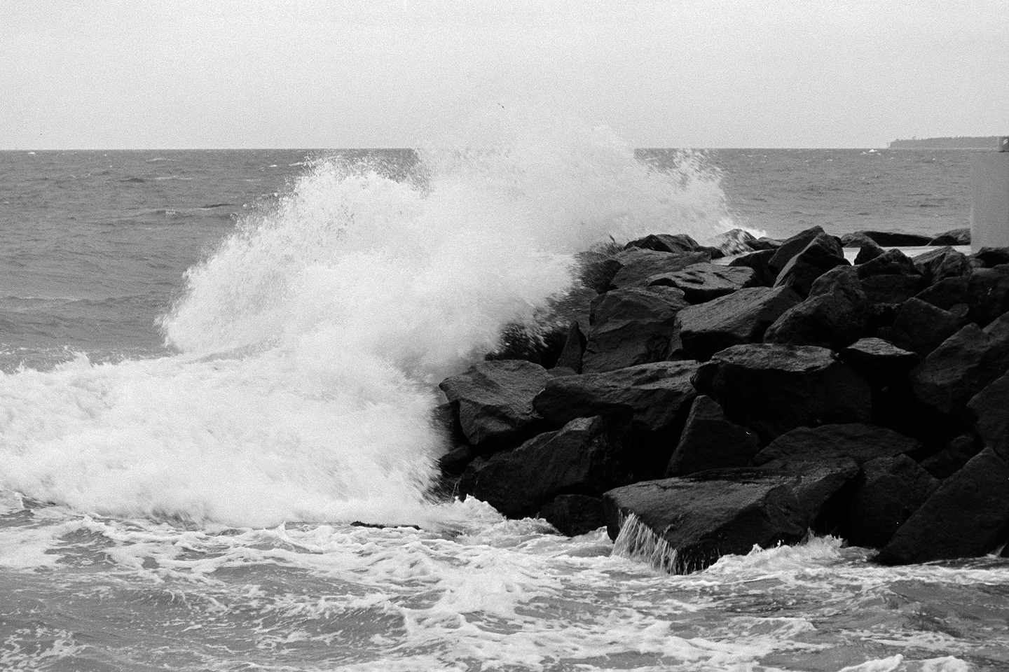 Grainy B&W shot of a powerful wave crashing against a dark rocky jetty under a moody, overcast sky.