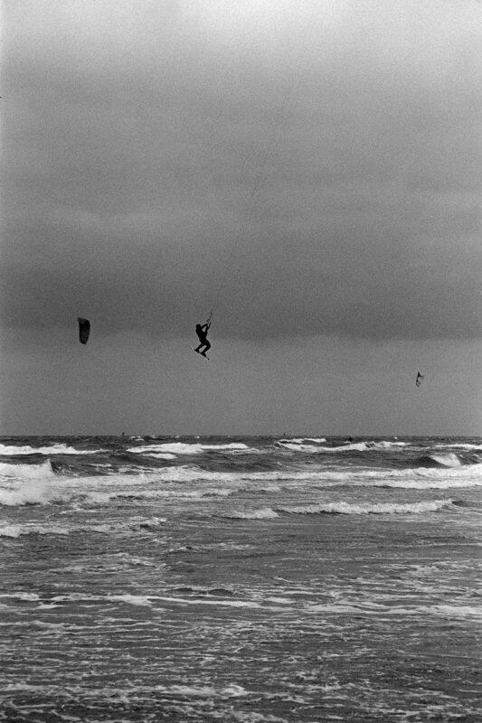 B&W film photo of a kitesurfer silhouetted against a vast, grainy sky above a choppy, dark sea.