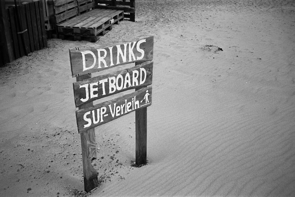 Grainy B&W close-up of a weathered wooden sign on the sand for drinks, jetboards, and SUP rentals.