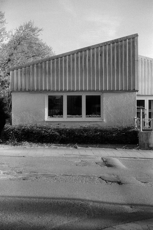 B&W film photo of a minimalist building with a slanted metal roof and a window reading "... täglich Mittagstisch ab 11.00 Uhr".