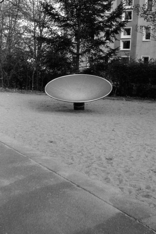 B&W film photo of a metal, dish-shaped playground spinner sitting empty on a sandy ground in front of a residential building.
