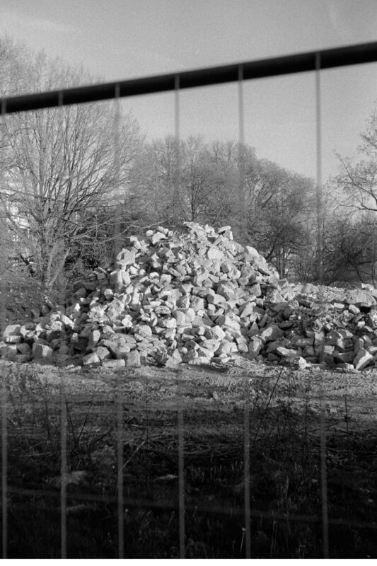 Grainy B&W shot of a large pile of crushed concrete and stones behind a construction fence, with bare trees in the background.