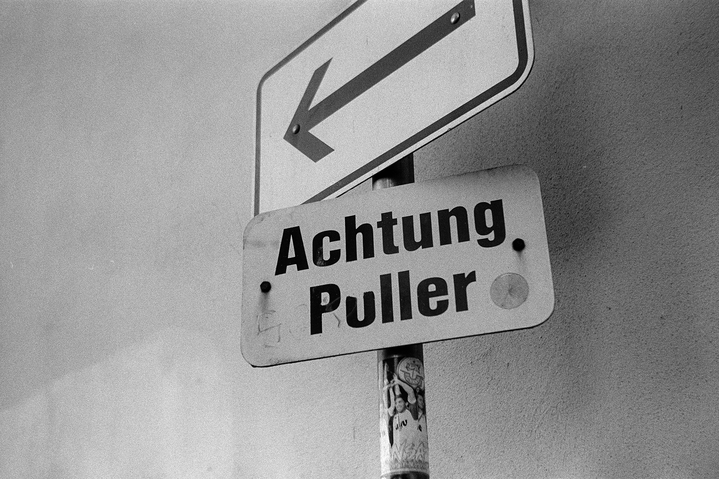 B&W close-up film photo of a street sign reading "Achtung Puller" beneath an arrow pointing to the left.