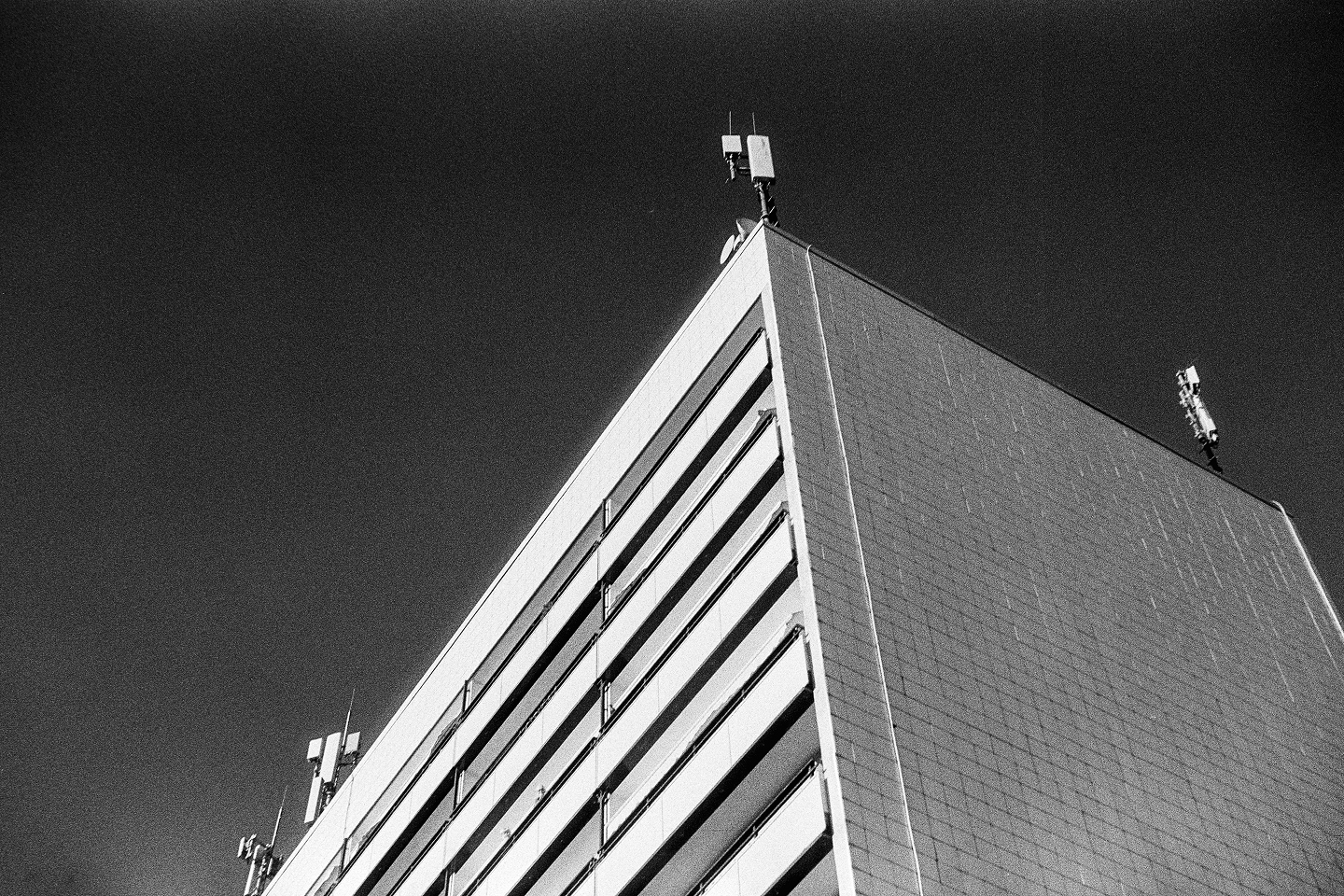 Low-angle B&W film photo of a high-rise building's sharp corner with antennas on top against a dark, grainy sky.