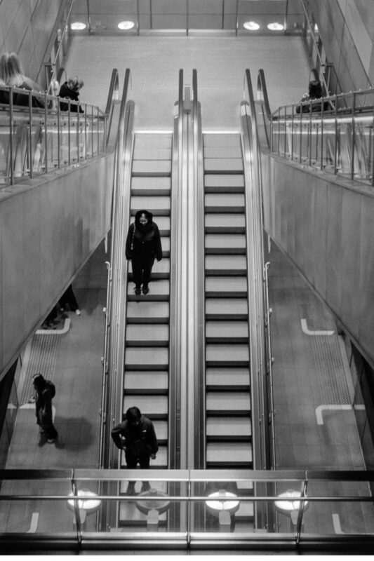 Symmetrical B&W film photo of commuters on long, parallel escalators inside a minimalist, industrial metro station.