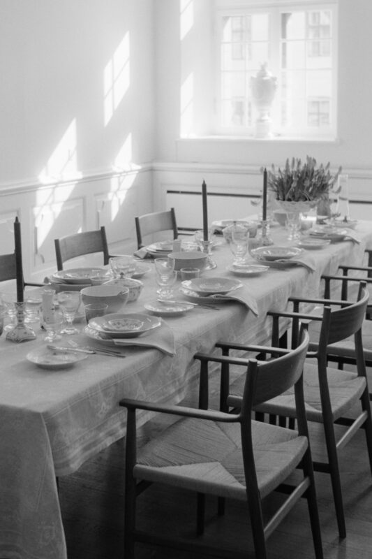 B&W film photo of an elegantly set dining table in a bright room, with sunlight creating geometric shadows on the wall.