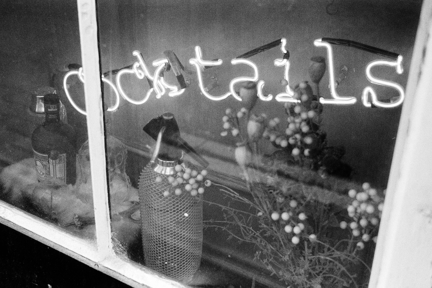 Grainy B&W close-up of a window display featuring a glowing neon "Cocktails" sign and a vintage glass siphon bottle.
