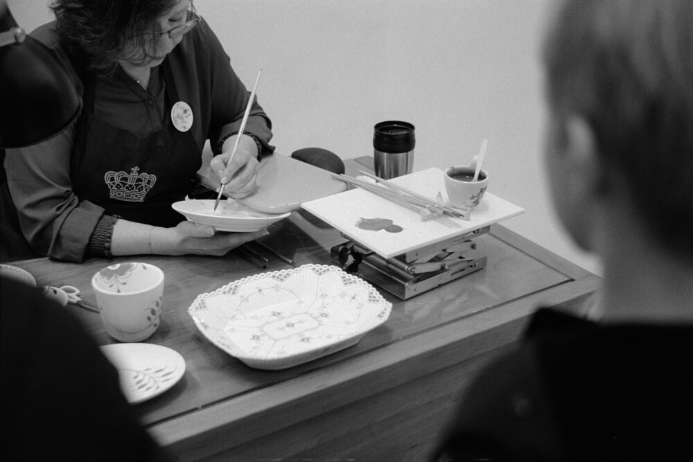 B&W film photo of a porcelain painter at Royal Copenhagen, meticulously detailing a plate with a fine brush.