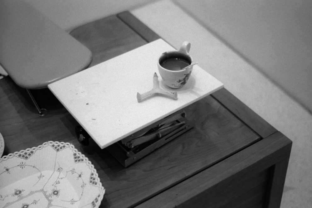 Grainy B&W shot of a workshop table with painting tools, a porcelain cup, and an intricate lace-edged plate.