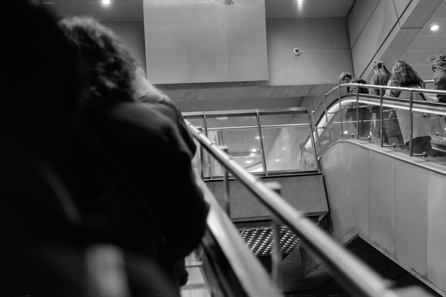 Low-angle B&W film shot capturing the dynamic movement of commuters riding an escalator inside a modern metro station.