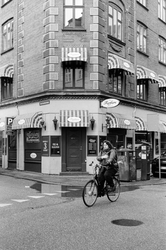 B&W film photo of a person riding a bicycle past a brick corner building with striped awnings and Danish signs in Copenhagen.