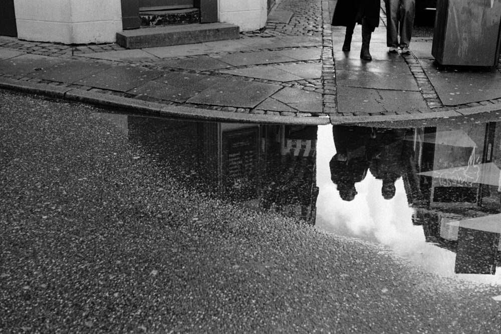 B&W close-up of a street puddle reflecting the silhouettes of two people walking on a wet cobblestone sidewalk.
