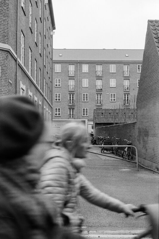 Grainy B&W shot with blurred people in the foreground, looking down a narrow alley toward a large multi-story apartment block.