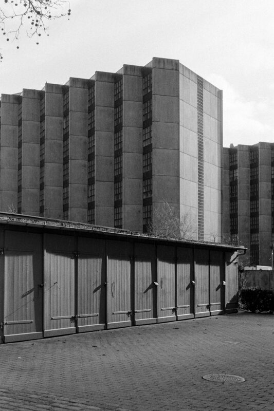 B&W film photo showing a row of low wooden garage doors in the foreground contrasted against a massive concrete Brutalist building.