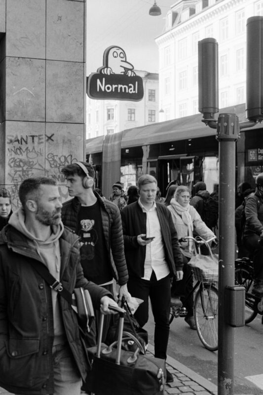 Grainy B&W street photo of pedestrians and a cyclist on a busy Copenhagen sidewalk near a store with a prominent "Normal" sign.