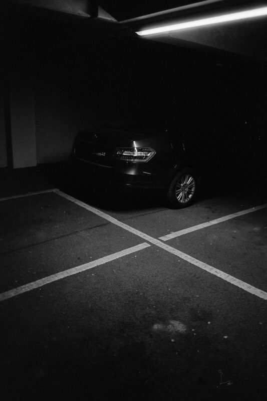 B&W film photo of a dark sedan parked in a dim garage, framed by stark white parking lines under an overhead light.