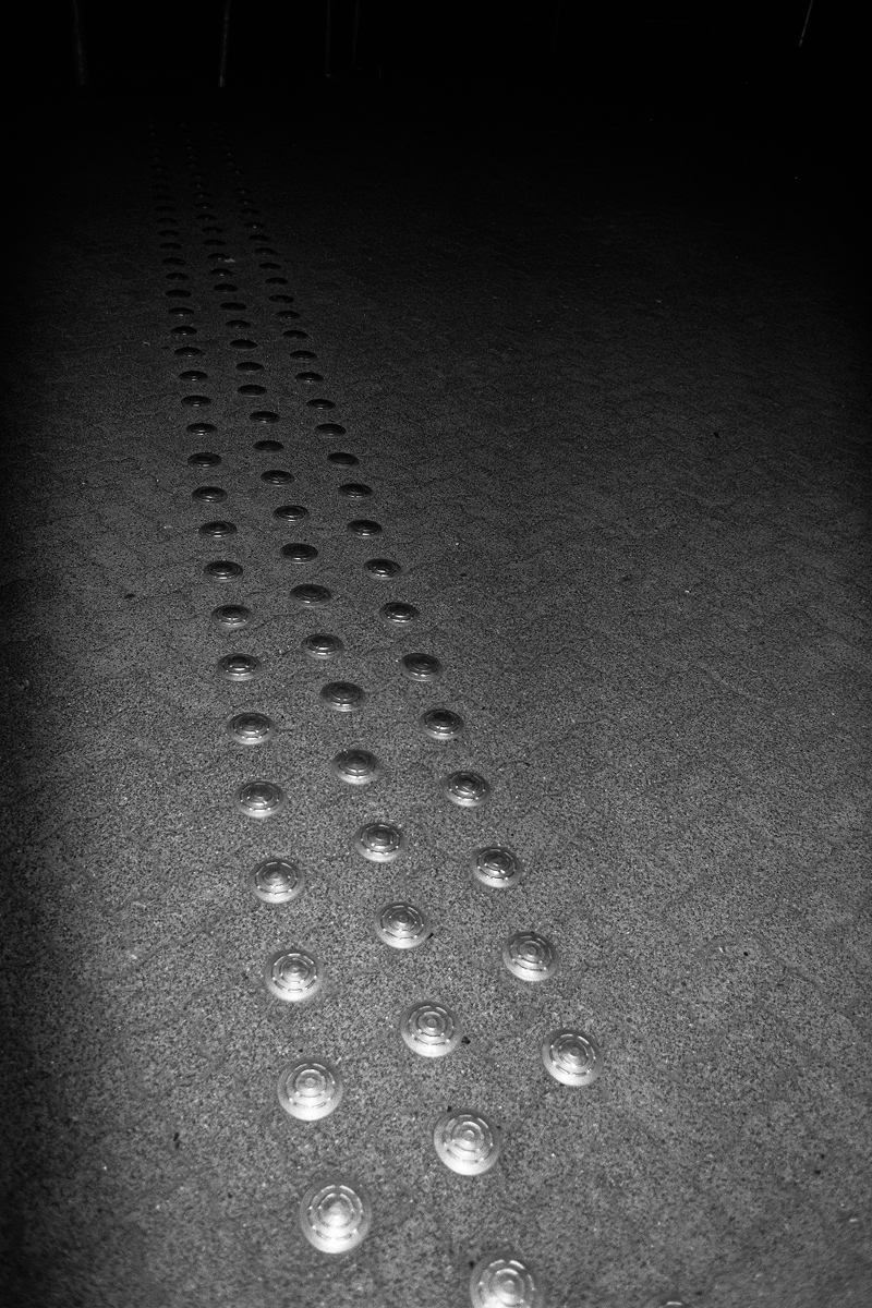 B&W flash photo showing a perspective view of metallic tactile paving studs on a grainy asphalt surface at night.