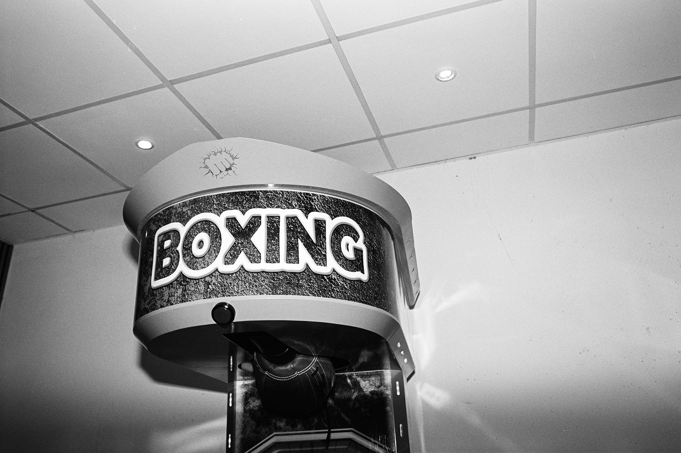 B&W flash close-up of an arcade boxing machine with bold "BOXING" signage under a grid ceiling at night.