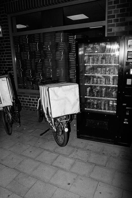 Grainy B&W flash photo of a delivery bicycle with a large insulated box parked next to a vending machine at night.