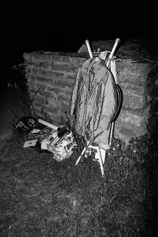 B&W flash shot of a winter coat on an ironing board left outside by a weathered stone wall at night.