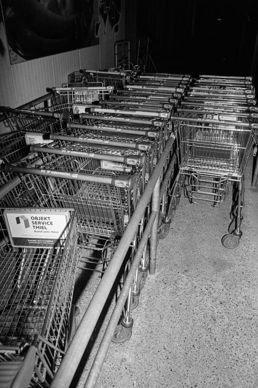 Grainy B&W flash photo tracking rows of nested shopping carts at night, with German advertising signs visible.
