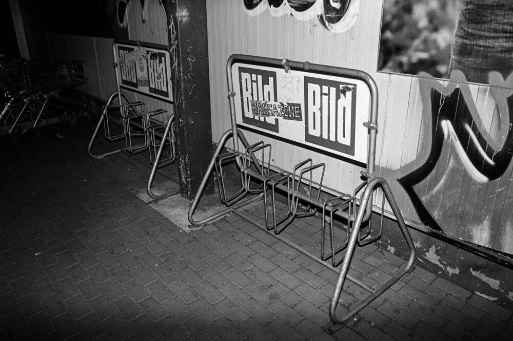 B&W flash shot of empty metal bicycle racks bearing German "Bild" and "Hansa Zone" stickers on a tiled ground at night.
