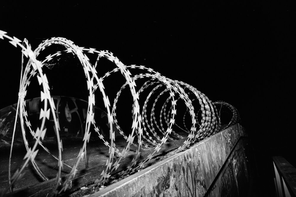 B&W flash photo capturing sharp coils of razor wire along a concrete wall against a black night sky with high contrast.