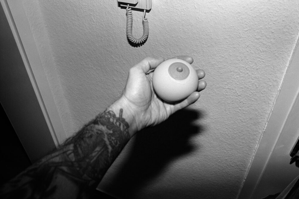 B&W film close-up of a tattooed arm holding a breast-shaped anti stress ball against a textured wall with a sharp night flash.