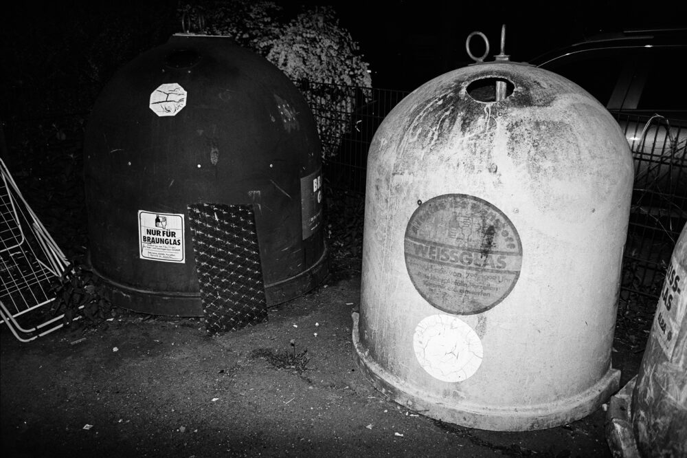 Grainy B&W flash photo of weathered public recycling igloos for brown and white glass on a dark street corner at night.