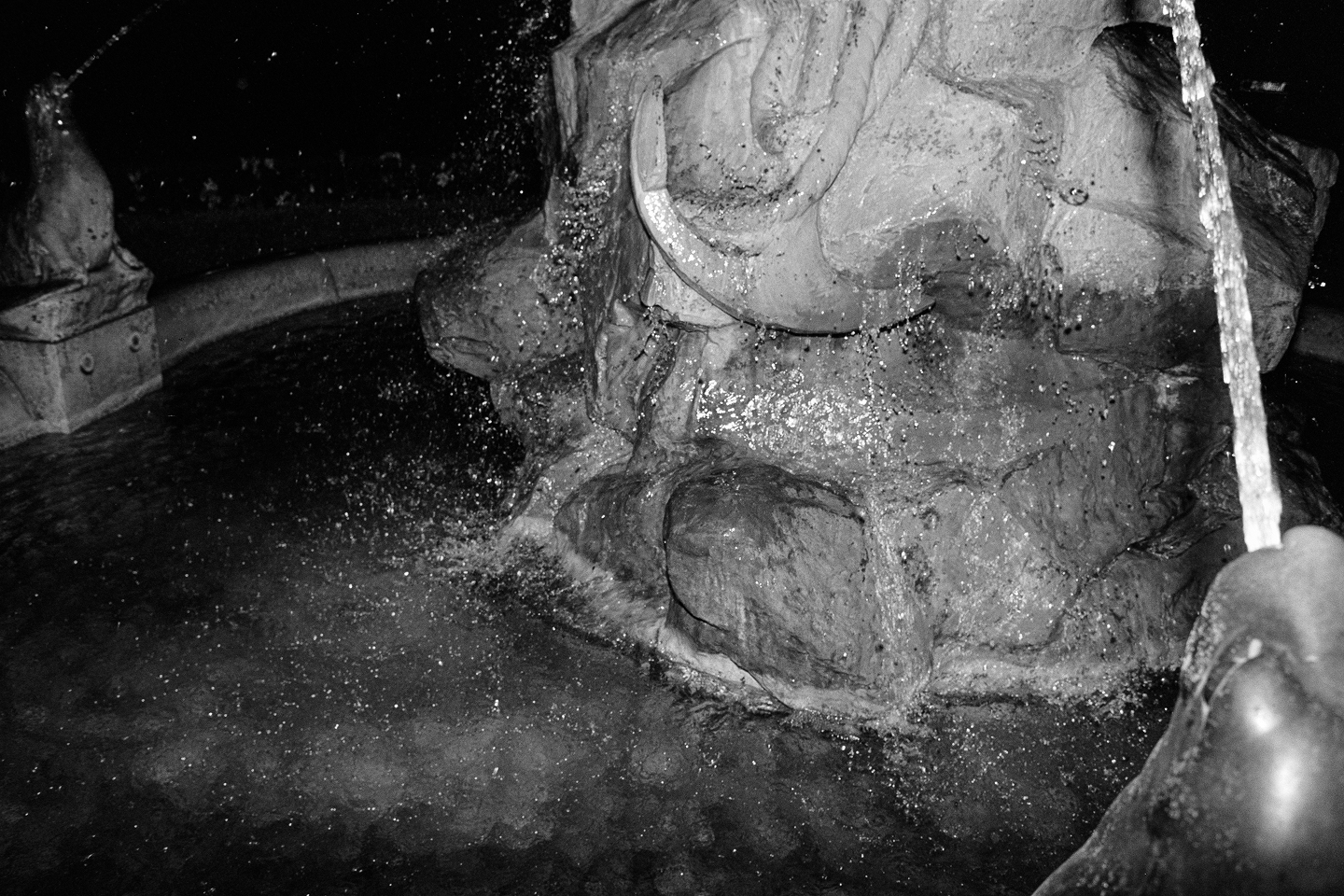 B&W film shot of water splashing over a textured stone fountain at night, illuminated by a harsh, dramatic camera flash.