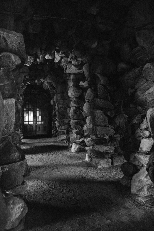 Grainy B&W shot looking through a dark, cavernous stone grotto tunnel toward a glass-paneled door with light shining through.