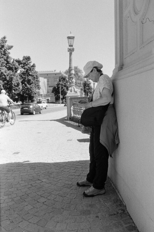 B&W film photo of a person in a cap leaning against a light-colored wall, looking at a device on a cobblestone street corner.