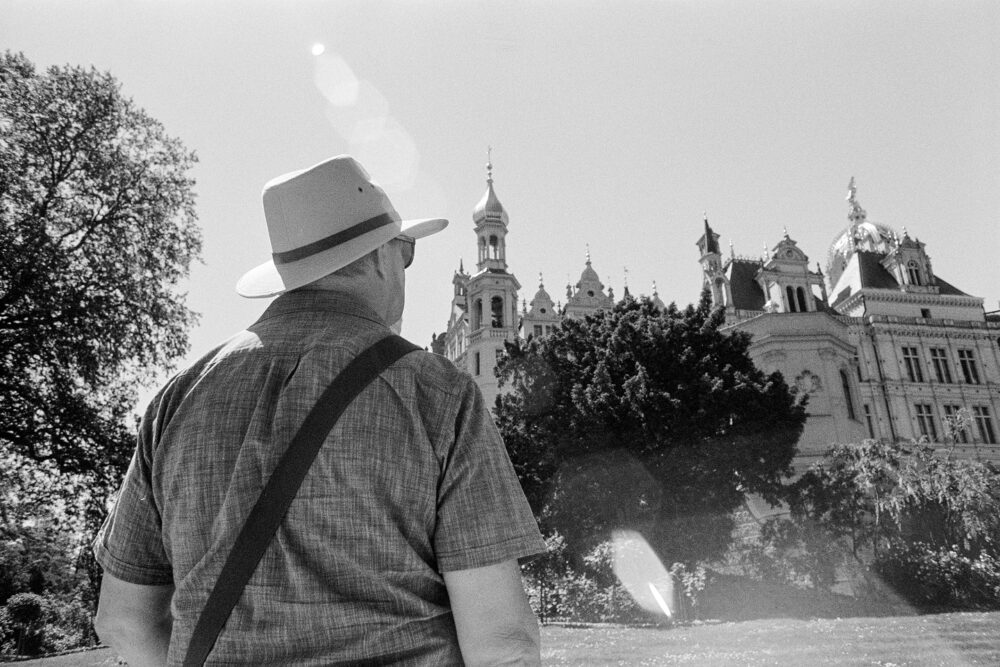 Grainy B&W photo from behind a man in a sun hat looking towards the ornate spires and towers of Schwerin Palace.