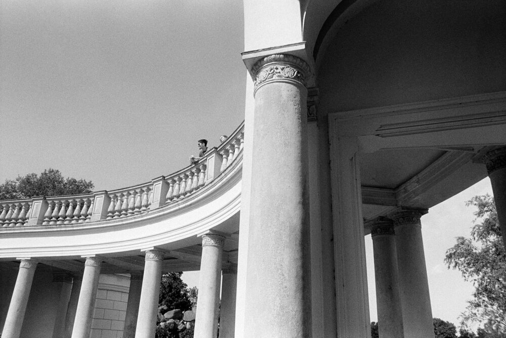 Low-angle B&W shot of a curved classical colonnade with stone pillars and balustrades under a clear, bright sky.