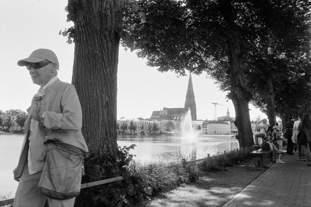 B&W film photo of a lakeside path with trees, featuring a distant church and fountain, with a blurred passerby in the foreground.