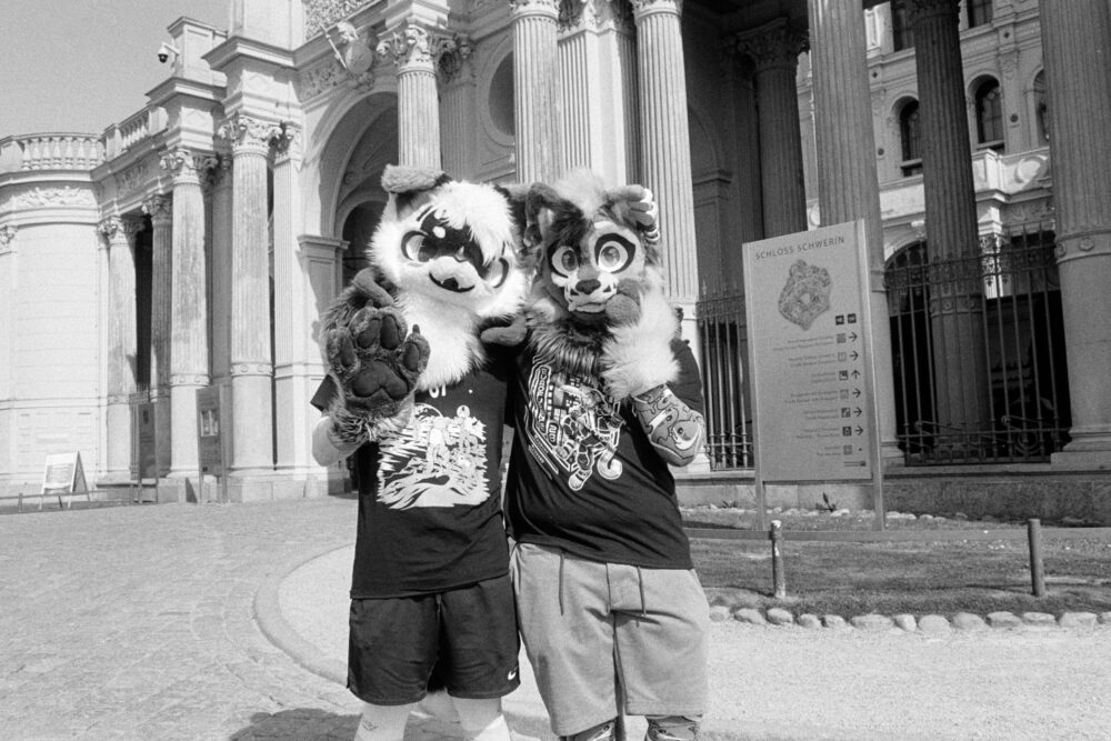 B&W photo of two people in furry animal costumes posing in front of the grand, pillared entrance of Schwerin Palace.
