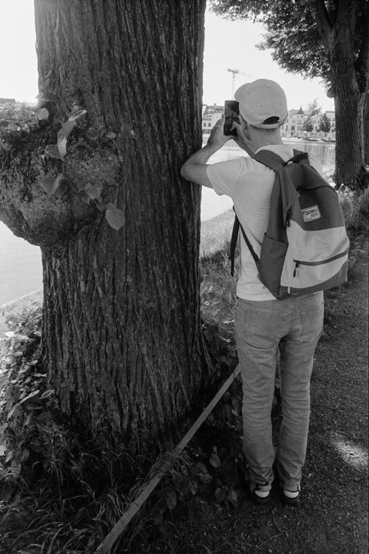 B&W shot from behind a person with a backpack taking a smartphone photo next to a large, textured tree trunk by the water.