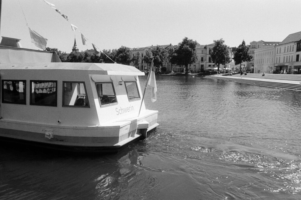 B&W film photo of a tour boat labeled "Schwerin" on the water, with city buildings and wide stone steps in the background.