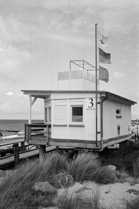 Grainy B&W film photo of a wooden lifeguard station marked with number 3, surrounded by wild dune grass on a cloudy beach.