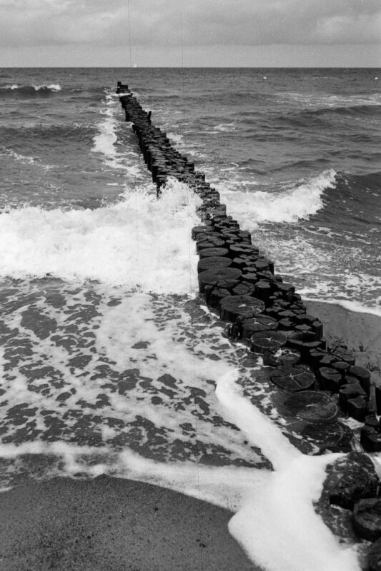 B&W film photo of a long wooden groyn stretching into the wavy Baltic Sea, showing textured waves and sea foam on the sand.